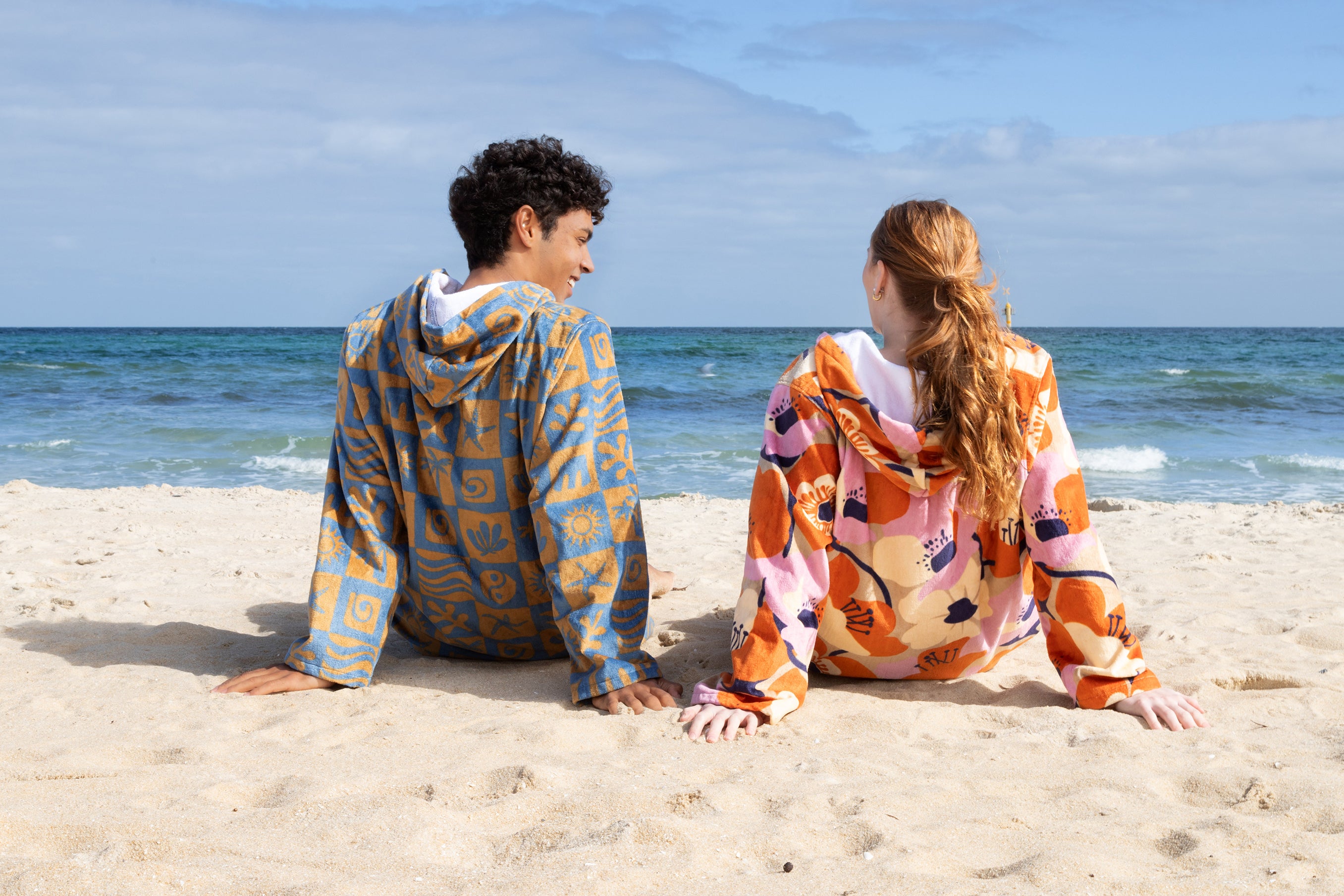 A couple wearing towel hoodies on the beach relaxing on the sand