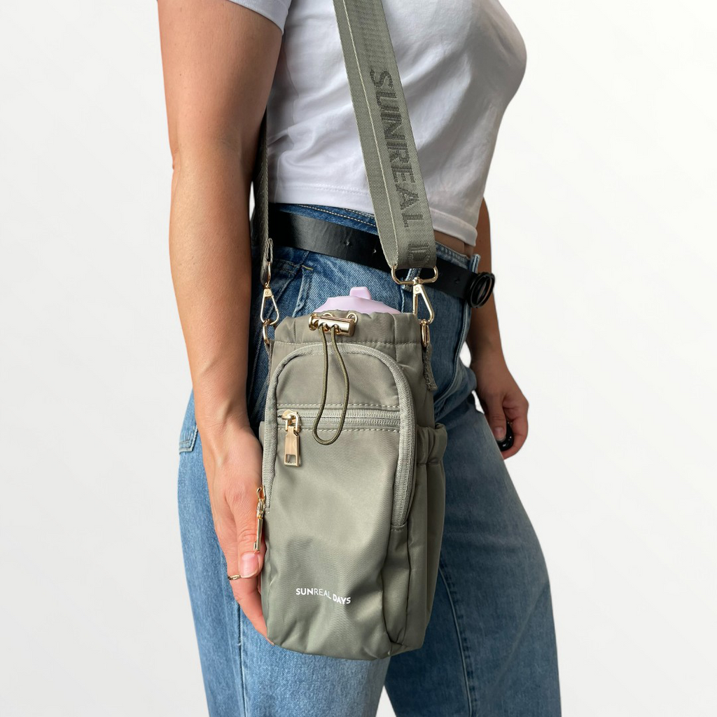 Lady carrying Khaki green drink bottle carry bag with gold accents and 'Sunreal Days' branding. Zipped pockets to hold phone and keys.