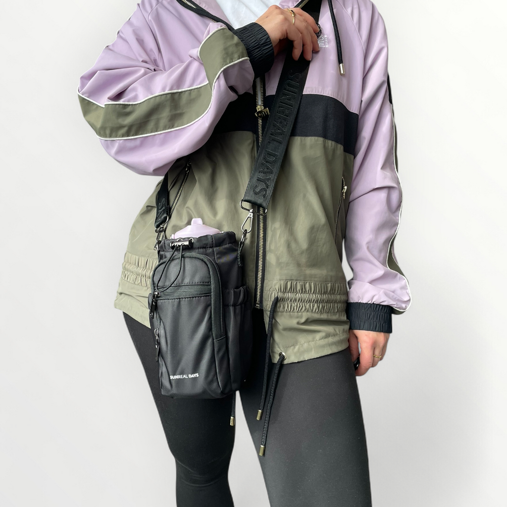 Lady carrying Black drink bottle bag with gun metal grey hardware and 'Sunreal Days' branding. Large zipped pockets to hold phone and keys.